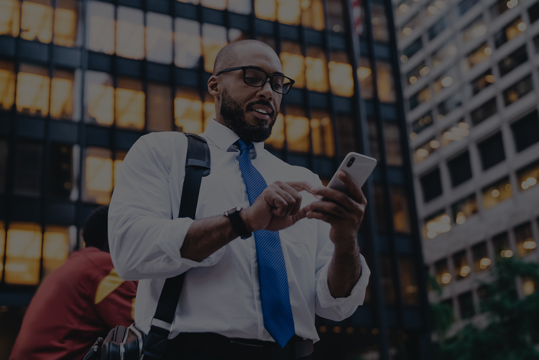 From below African American bald bearded employee with glasses messaging on white smartphone while standing in front of modern office buildings